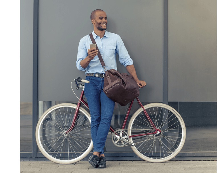 Full length portrait of handsome young Afro American man in casual clothes using phone, looking away and smiling while leaning on his bike, standing outdoors